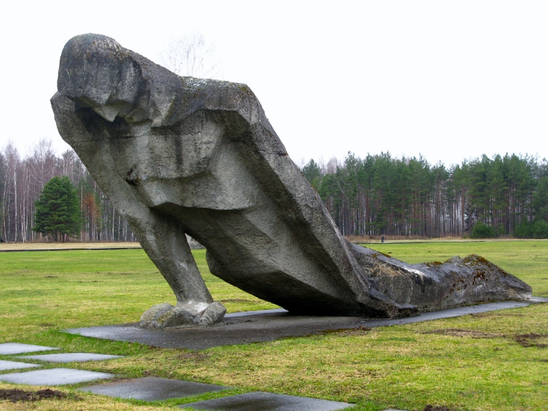 a massive stone sculpture of a man lying on the ground, his right arm ended to prop up his torso. His feet drag behind him. It's set on stone in a grass field with a row of trees at the back against a grey sky
