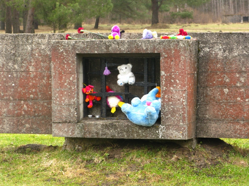 A metal grate in a long, low, brick wall in which soft toys hang. Teddy bears - red, white, blue, purple all left as memorials