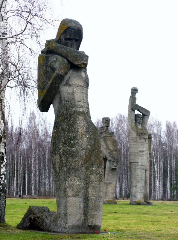 Three mammoth stone statues. In the foregroun, a keeling woman with her arm hugging her chest. In the back ground, two men, one with his right arm extended over his head, with his left arm grasping his elbow. The other simply stands. In the back a line of leafless trees stand against a grey sky in sharp contrast to the green grass