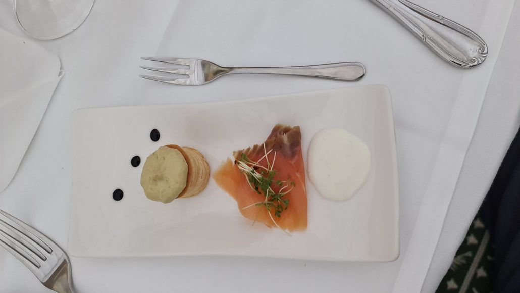 A white rectangular plate flanked by two forks on a white table cloth. On the plate is an amuse bouché of a tiny puff pastry topped with pumkpin cream and some slices of smoke salmon topped with watercress