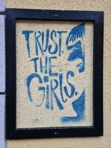 Blue graffiti on white in a black frame: Trust the girls