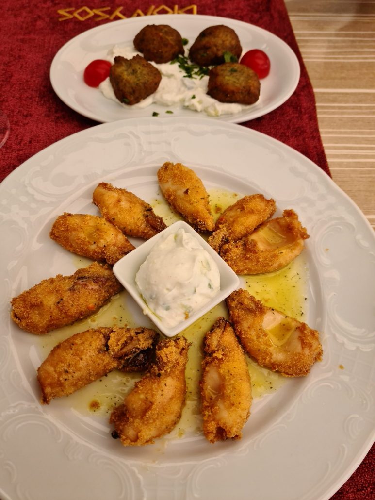 Plate of 9 piece of calamari fried in garlic butter with some cucumber yoghurt in a bowl in the middle. To the left another plate of 4 fried zucchini cakes