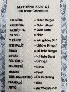 Paper napkin edged in blue designs with common Greek phrases translated into german