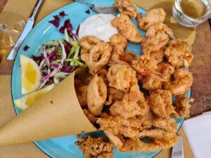 A brown paper cone from which spills fried calamari and shrimp set on a blue plate on a bed of purple shredded cabbage