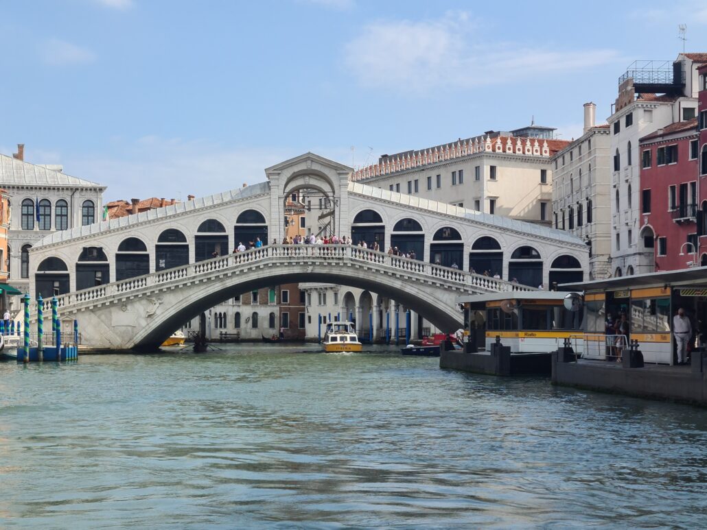 The Rialto bridge in Venice - an inverted V bridge with six arches on either side and a large arch in the centre. A yellow vaporetta passes underneath