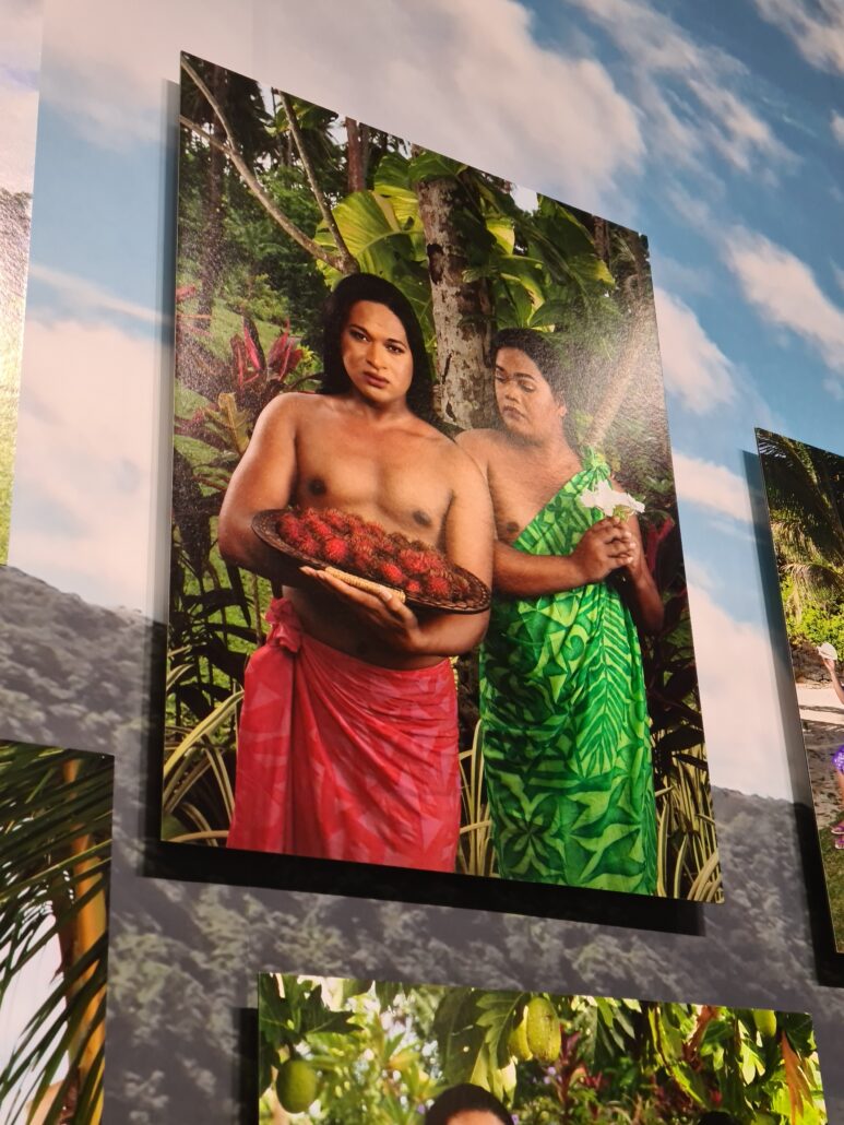 Portrait of two Samoan Fa‘afafine. One barebreasted waiting a red sarong around their waist is holding aholding a platter of red fruit. The other looks over their shoulder at the fruit and wears a green sarong pinned over their shoulder. Both as set agains a rainforest background. The painting is on a wall painted with a blue cloudy sky. The end of another paint is in shot to the right and the top of a third pops up below. . 
