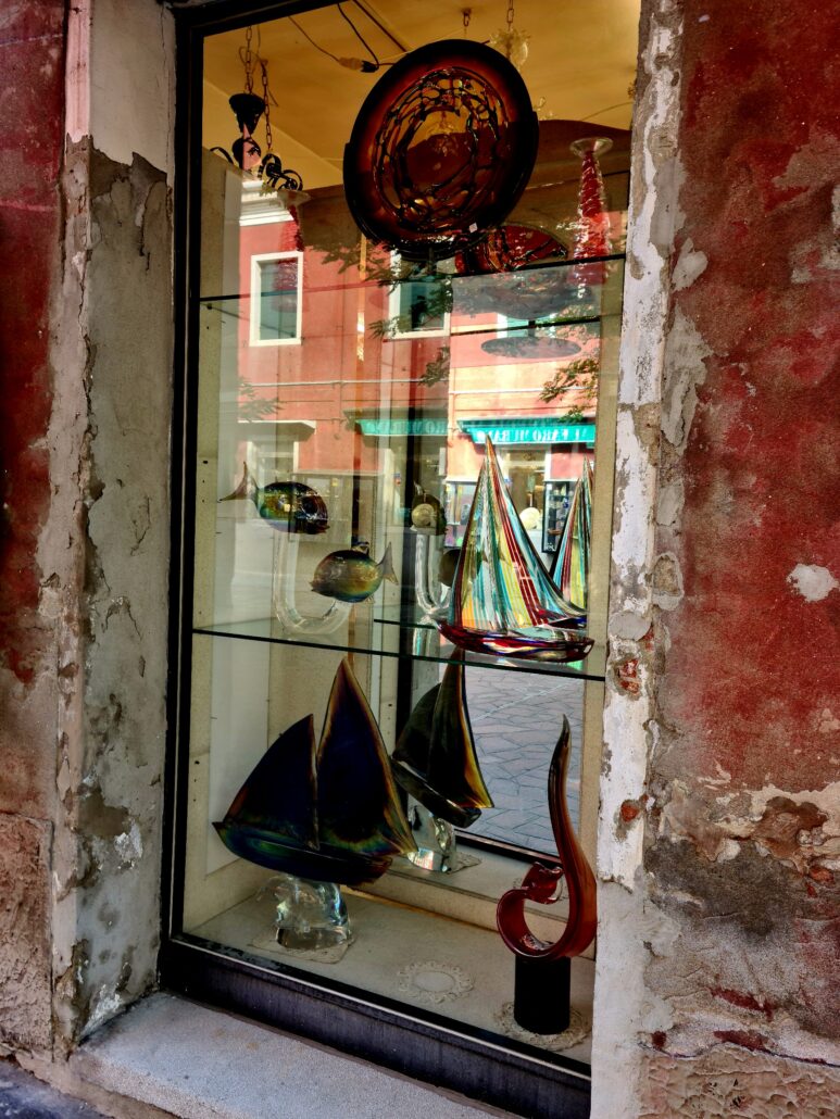 shop window showing colourful boats made from Murano glass - red building across the road reflected in the window