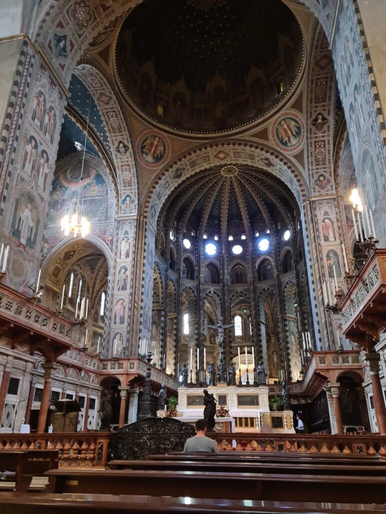 The elaborate interior of the Basilica of St Anthony of Padua. A lone man sits in a pew praying in front of the altar. The walls and arches are covered in frescoes. 