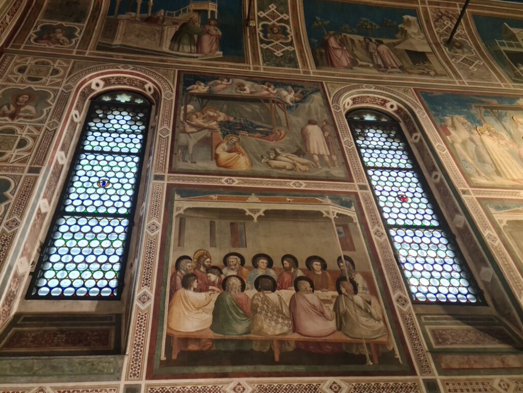 Two tall arched windows divide a wall of frescoes depicting christ's life. In the foreground is a painting of the last supper. Above it is a nativity scene. 