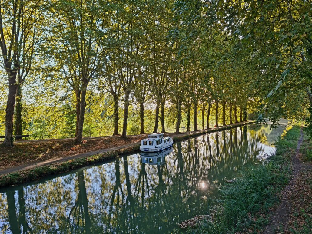 White barge moored at the side of a canal. Trees lining the left side are reflected in the still water