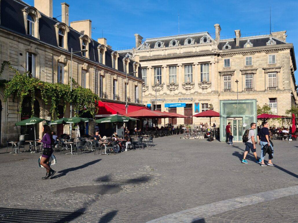 One corner of an old square with a three-storey building marked with the words Universite Bordeaux to the right. Rend and green umbrellas market the outdoor cafés 