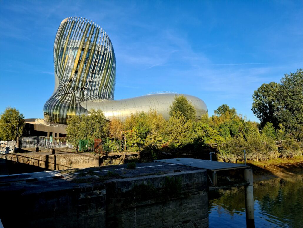 Steel building designed to look like a wine decanter set against a blue sky with leafy gree trees in the mid ground and water in the foreground