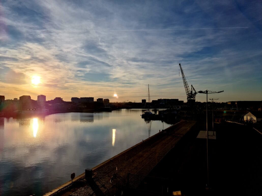 Sun setting over water at a dock with tall buildings and a large crane in the distance