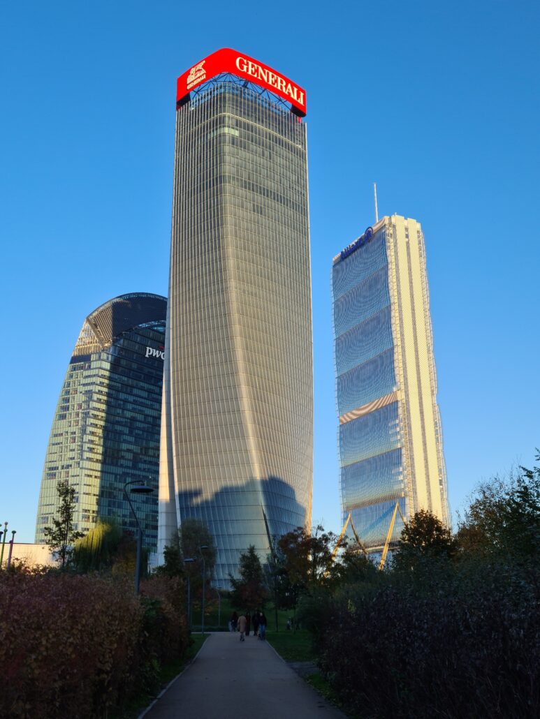 Three skyscrapers set against a cloudless blue sky with a tree-line path in the foreground. All are labelled. Left - PWC, Middle, Generali, Right - Allianz. 