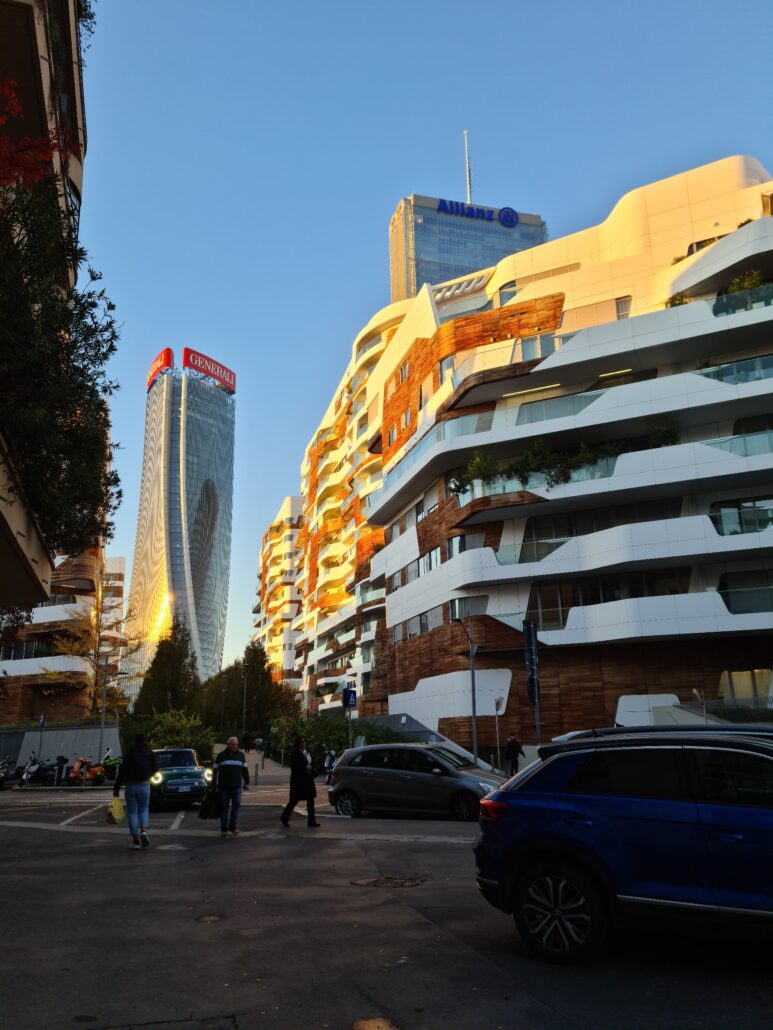 Street scene with a curved skyskraper with a red sign on top. White letters spell Generali. To the right is a block of flats - the sun has painted the tops of them a bright yellow while those in the shade are stark white. People walk in the street and cars drive by. 