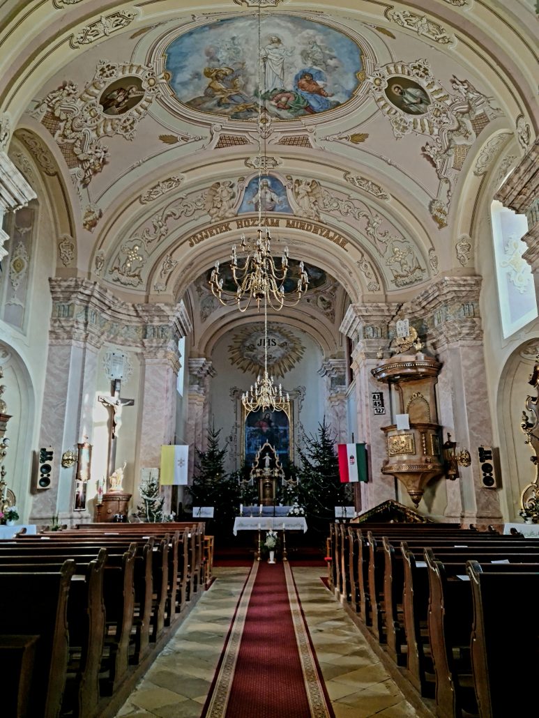 Interior of a church with frescoed ceilings and ornate chandeliers. A red carpet on gold tile marks the centre with rows of wooden pews to either side. 