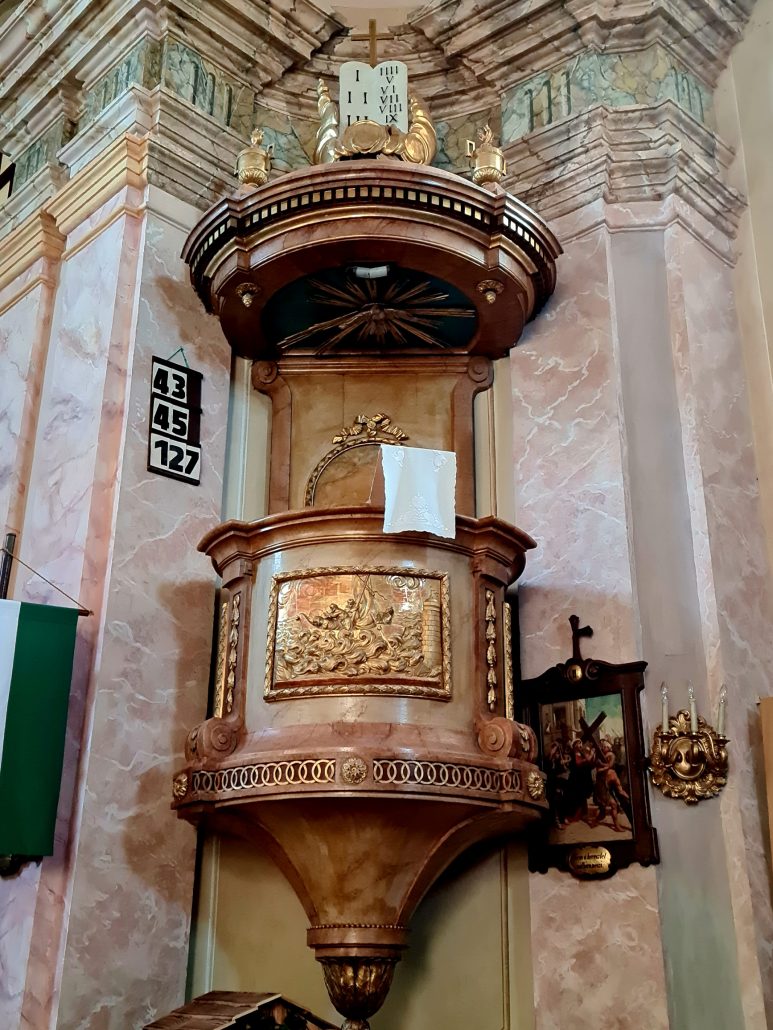 A church pulpit in gold and copper set against a pink and white marble wall with green trim at the top. To the left the numbers 43, 45, and 127 are set black on white showing the hymn numbers to be sung. To the lower right is a station of the cross. To the left we can see the green and white stripes of a flag. 