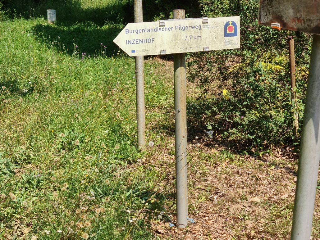 signpost pointing the way to Inzenhof 2,7 km. Burgenlandsicher Pilgerweg. With the symbol of a church - a round red dot in a white ellipsoid topped with a white cross all set into a blue ellipsoid. 
