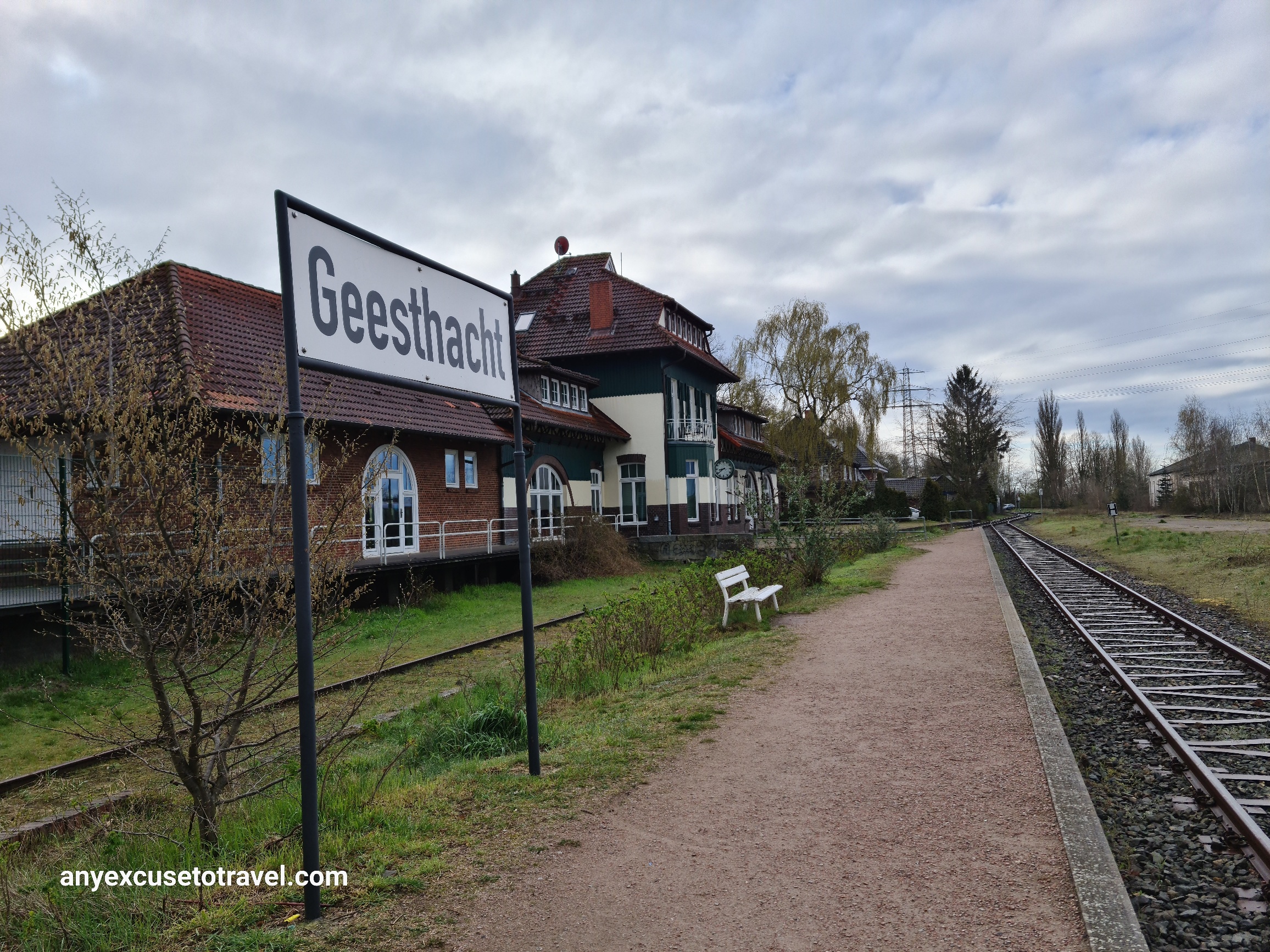 Sign - black letters spell Geesthacht on a white background - beside a railway track with a redbricked train station in the background to the left