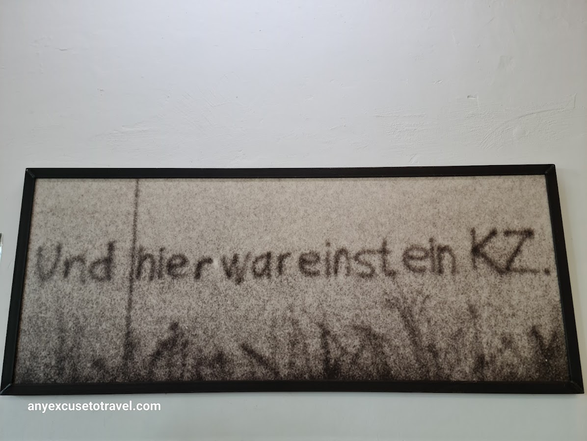 Framed photo on a while wall. The photo is of a wall onwhich is written Und hier war einst ein KZ (and here was once a concentration camp)