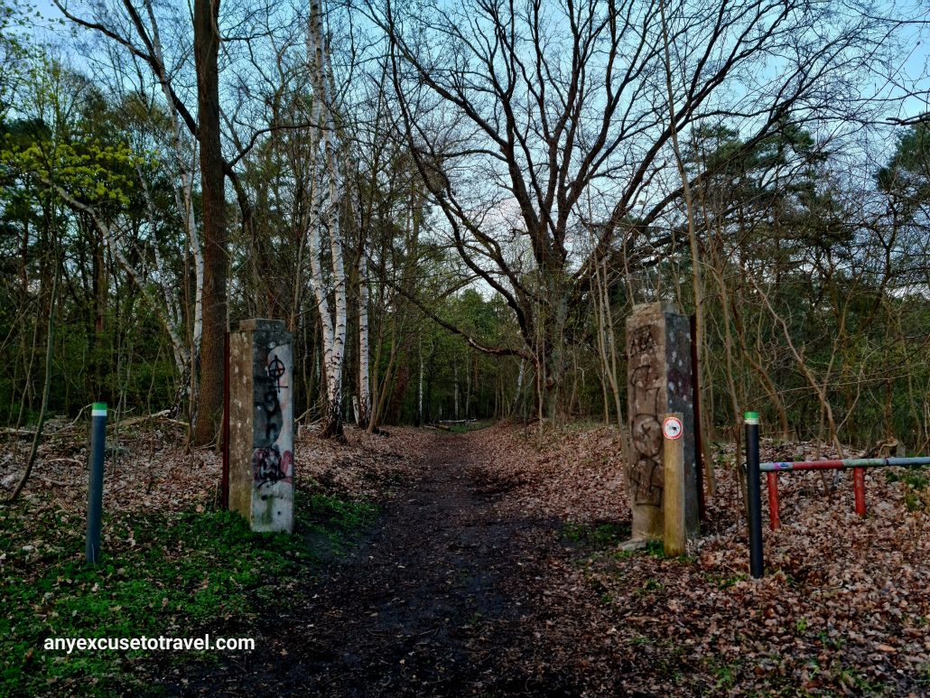 Forest path with two stone pillars on either side and a red and green barrier to the right. The pillars have old Roman guards painted on them. 