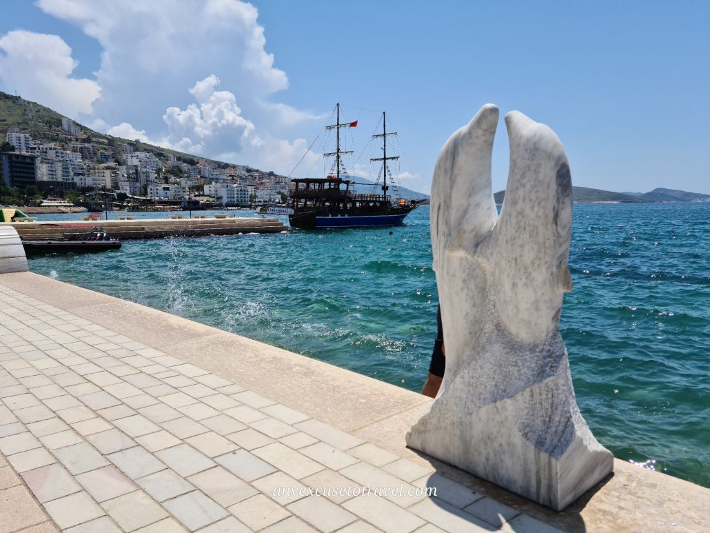 Granite statue of two dolphins dancing on a paved promenade beside clear blue waters. A pirate ship is docked in the background and buildings creep up the side of a hill. 