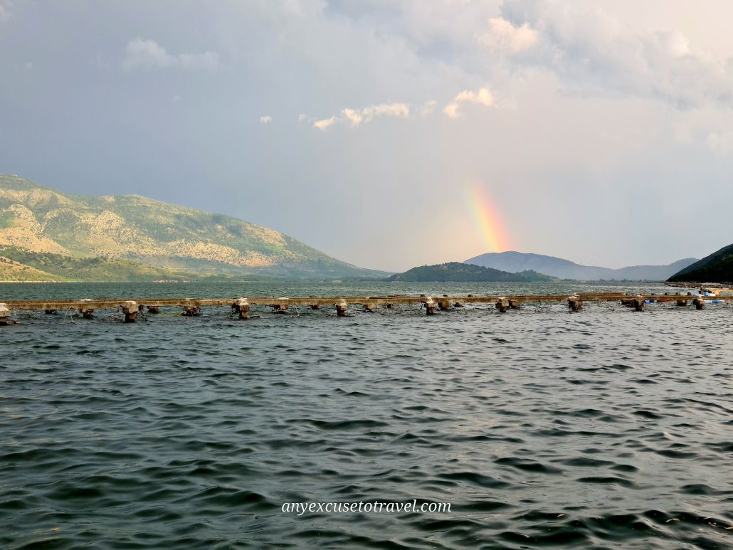 A row of concrete overhangs riding the surface of a lake (a mussel farm) a rainbow starts from the top of a mountain in the backgrund