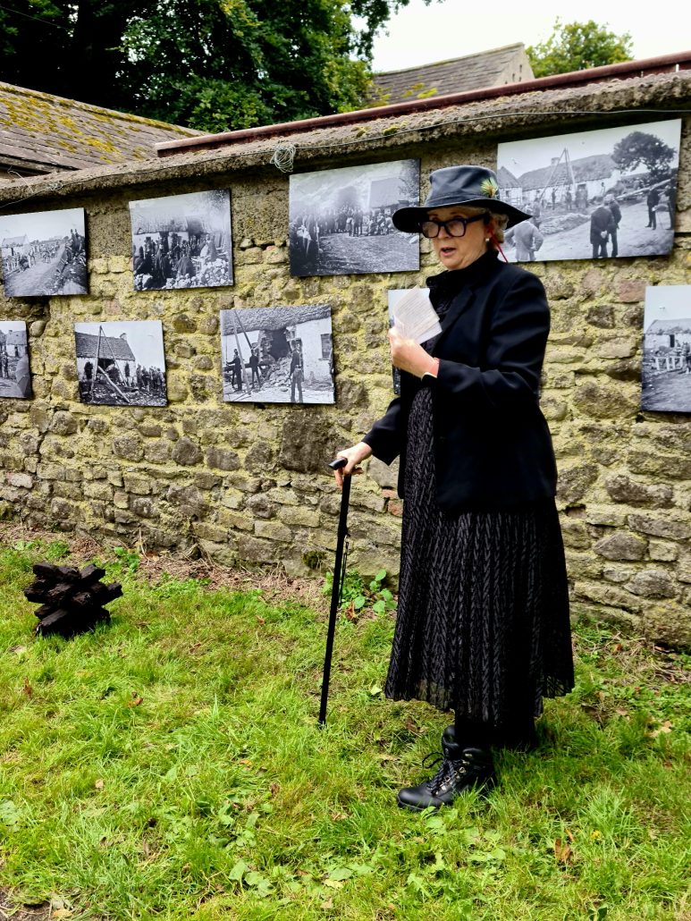 Yellow stone wall with two rows of black-and-white photos showing evictions from the 1880s in Ireland. A women in a black hat, black glasses, black pleated skirt and black jacket with black laced books stands with a walking stick holding a white card. 