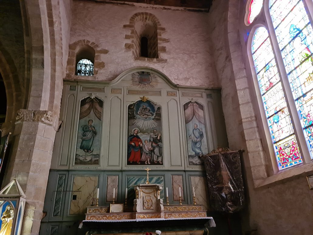 Church niche with a wooden altar - three panels - left and right are men in robes, middle is the holy spirit descending on the apostles. A crucifix below on an altar is flanked by three white candles on ither side - stained glass window to the right