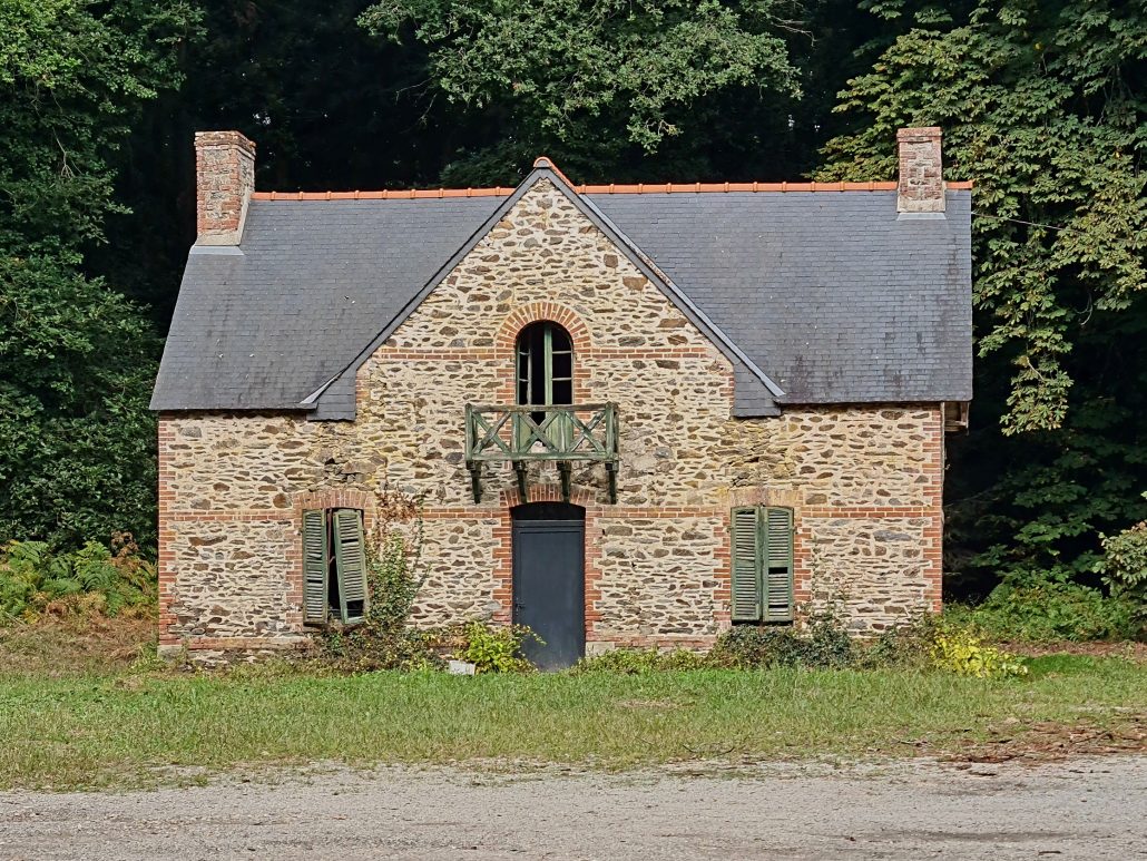 Stone house - a chimney at either end - two green shuttered windows flank a grey door above which is a a broken green wooden balcony in front of a partially open window. 