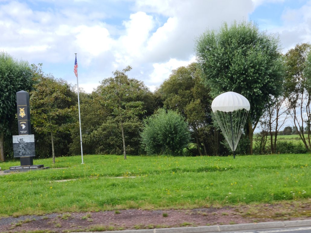 Between the grass verge and the treeline is a monumen tof some sort, a flag pole with a limp American flag, and a small white parachute. 