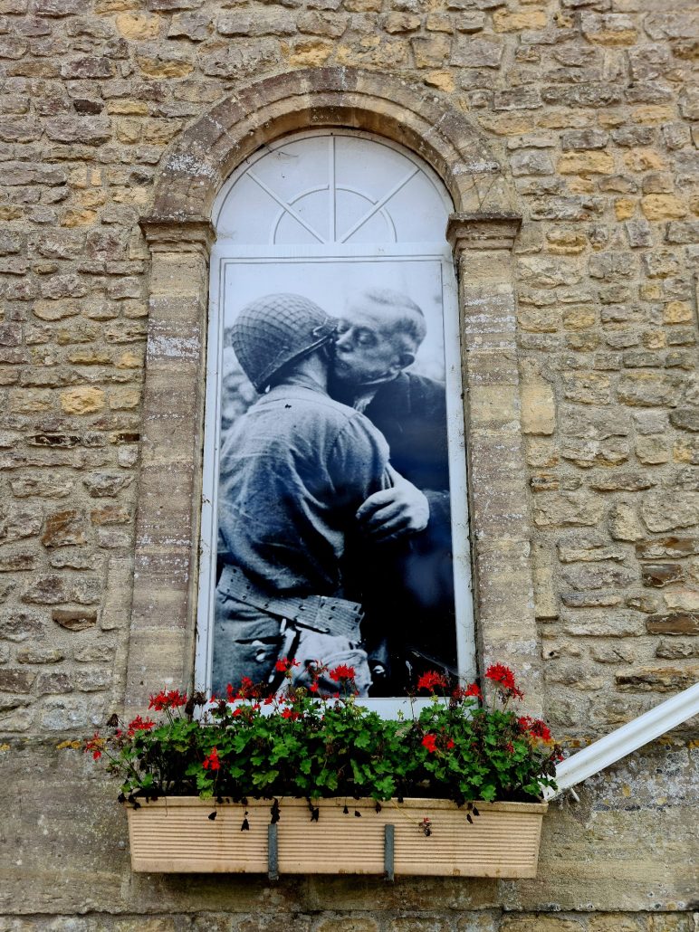 Arched niche in a yellow stone wall has an inset of a blown-up black and white photo of a man kissing a solider - a windowbox with red flowers sits underneath