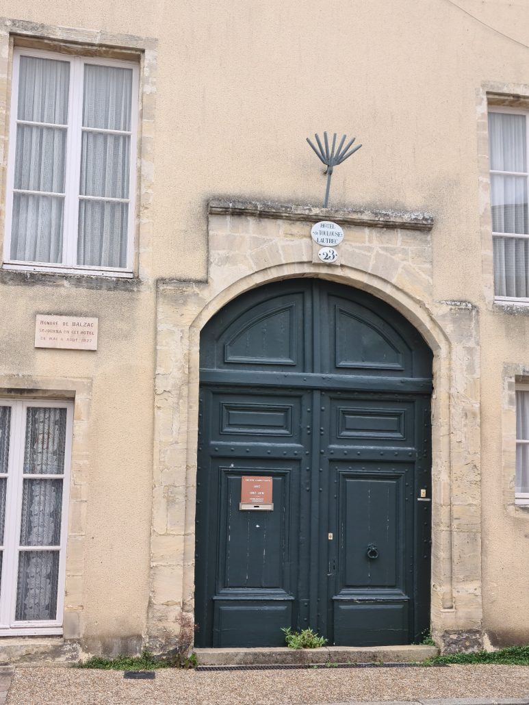Portion of a yellow stone building with a green arched door on which is a red plaque with the words ANCIES COMBATTANTS UNC UNC-AFN PERMANENCE written in white. Above the door are two more signs, in white. One says: Hotel de Toulouse de Laugrec and the second has the number 23. To the left of the door between the two six-paned white windows is another: Honoré de BALZAC sejourna en cet hotel de mai a aout 1822. Weeds grow in the ground in front of the door. 