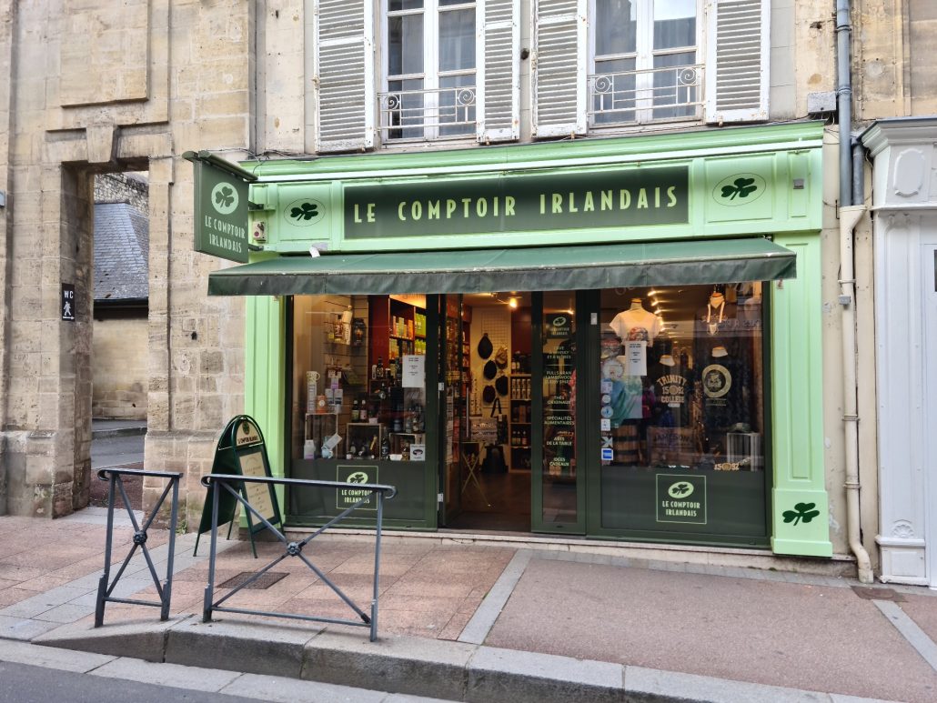 Green shopfront with the words LE COMPTOIR IRLANDAIS written in pale green on dark green with a shamrock on either side. Shop window is home to t-shirts, hands, and other knickknacks