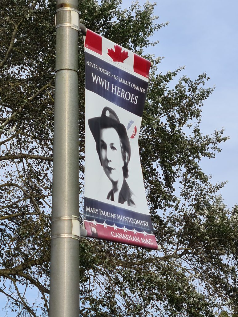 Flagpole with a banner - top canadian maple leaf in red on white - middle - NEVER FORGET/NE JAMAIS OUBLIER WWII HEROES - then a headshot of a woman in military uniform. Underneath: MARY PAULINE MONTGOMERY CANADIAN MAC - behind the banner is a green tree against a grey-blue sky