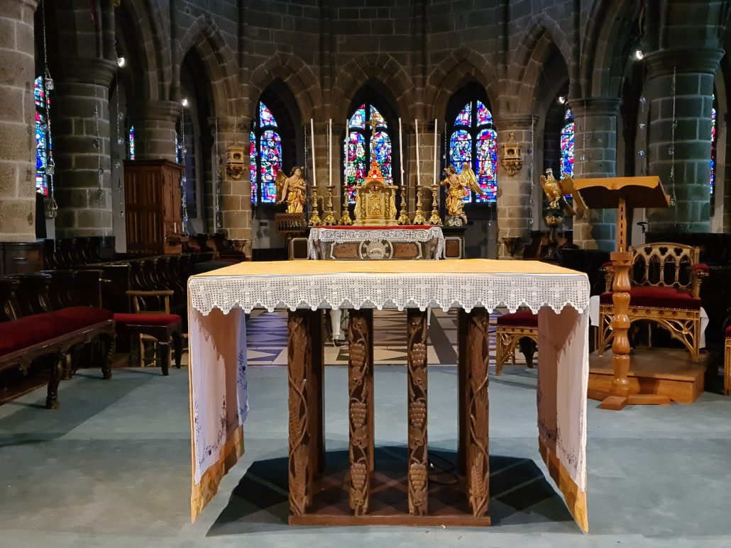 Church altar draped in a laced tablecloth. Wooden lectern to the right with a carved wooden chair with a red velvet seat behind it. Another atlar behind has the tablernacle with three tall white candles on either side. These are flacnked by statues of angles. Stained glass windows visible at the back and sides. 