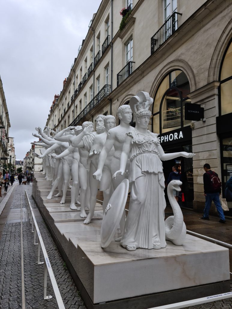Street sculpture of a line of Greek statues standing in the middle of a pedestrianised shopping street. They're standing one behind the other, most likely characters from Greek mythology. They're all in white. 