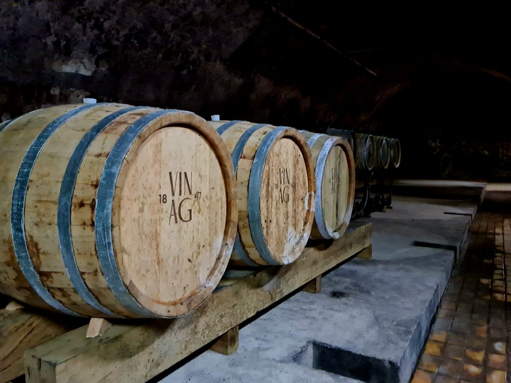 Three wooden wine barrels bearing the notation 18 VIN 47 AG Barrels sit on a wooden stand. There are more in the background. 