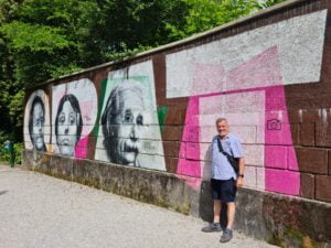 Himself (bearded grey-haired man in shorts and t-shirt wearing a black bag, messenger style, standing in front of a self-portrait background painted on a wall of portraits. Beside his is a portrait of Einstein