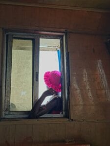 Side profile of an African woman wearing a cerise pink hat as she leans against an open window