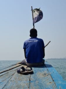 Sierra Leonian boy sits on the bow of a fishboat with is back to us - on his blue polo shirt is written BOAT MAN. The country's flag flied overhead - and his sandals are on deck behind him