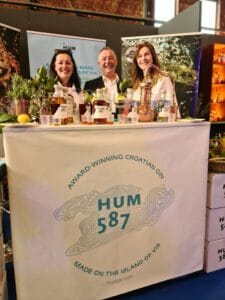 A man flanked by two women standing behind a exhibition stand. Table covered in a white cloth with blue writing - Award-winning Croatian Gin - HUM 58 -