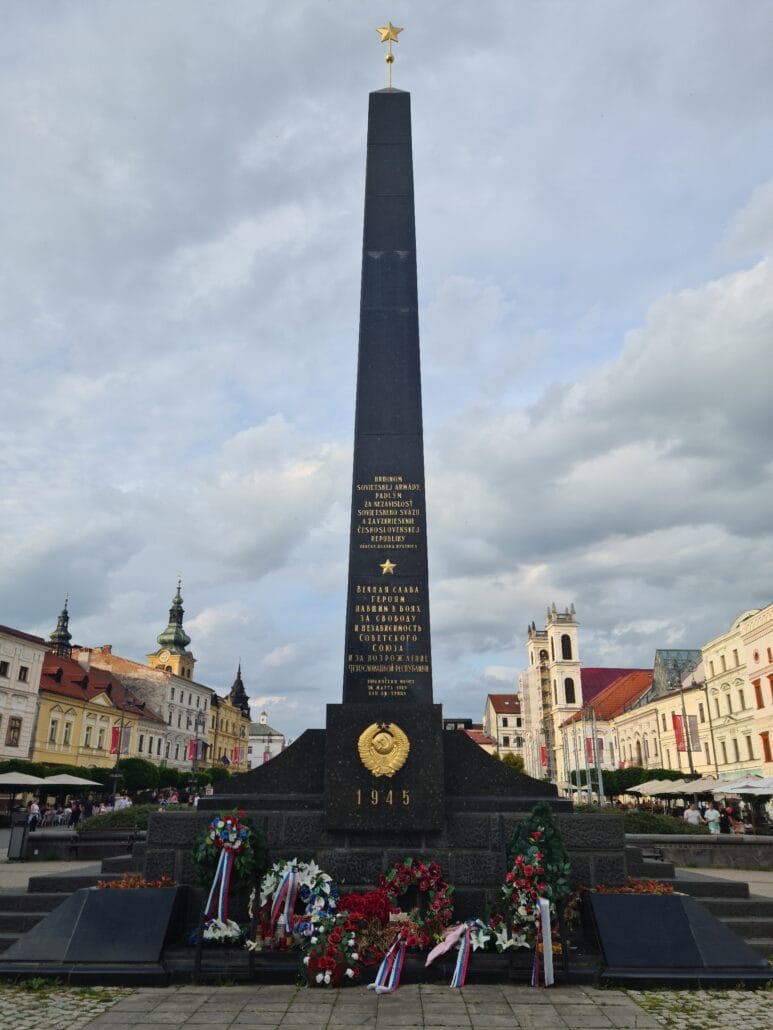 Black marble obelisk with the gold start on top. Gold inscription marks it as a monument the Russian liberators of 1944. Wreaths are scattered at the base. In the background is a square lined with baroque/gothic/renaissance buildings