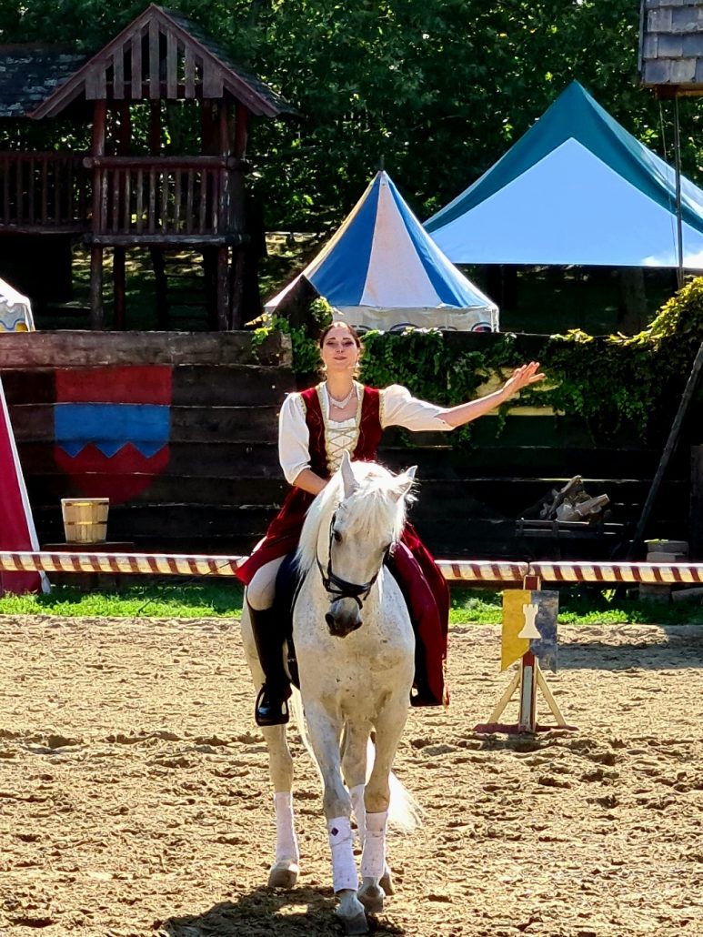 Woman in medieval dress on a white horse holding out her arm as if about to bow