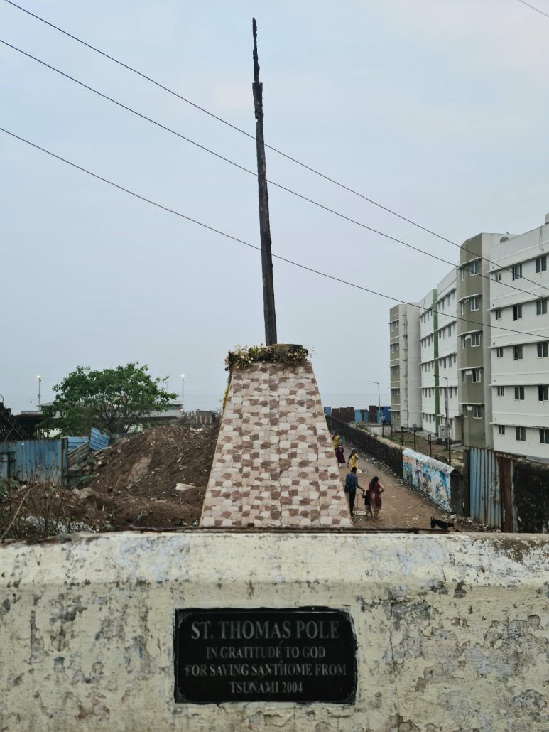 A tall post set in a stone base with the inscription on the wall reading St Thomas Pole In Gratitude to God for saving Santhome from tsunami 2001 - tall 5-floor apartment buildings to the right - electricity lines passing overhead - people walking on a dirt road towards the water (not visible)