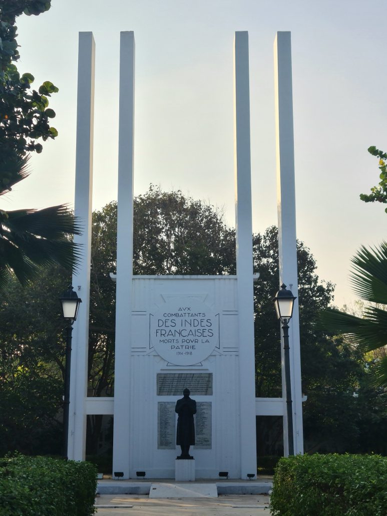 White war memorial consisting of four marble pillars with a front-facing bronze plaque containing the names of fallen soldiers. The central statue of of a French soldier resting on his rifle