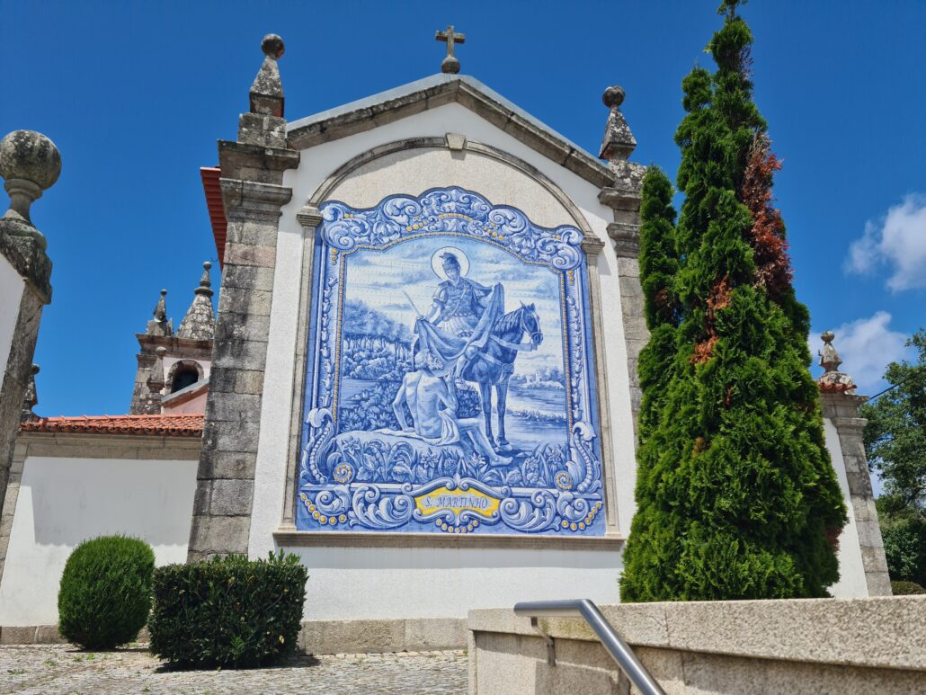 Back wall of a church with traditional blue-and-white tile depicting the famous image St Martin shredding his cloak in half to share with a poor man he encountered at the city gate of Amiens. A large fir tree grows on the right - brown bits show that the tree is diseased