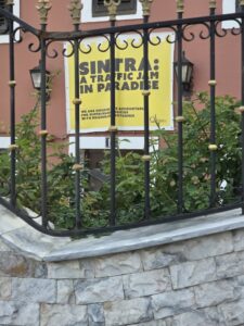 Yellow sign with black writing behind a railing on a red house. Sign reads: Sintra: a traffic jam in paradise. We are holding (?)s accountable for sustainable policies with measurable outcomes