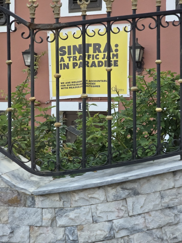 Yellow sign with black writing behind a railing on a red house. Sign reads: Sintra: a traffic jam in paradise. We are holding (?)s accountable for sustainable policies with measurable outcomes