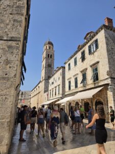 A sunlit historic stone street lined with traditional buildings, green-shuttered windows, and awnings, with a tall stone bell tower rising in the center under a clear blue sky. Tourists walk through the busy pedestrian street.